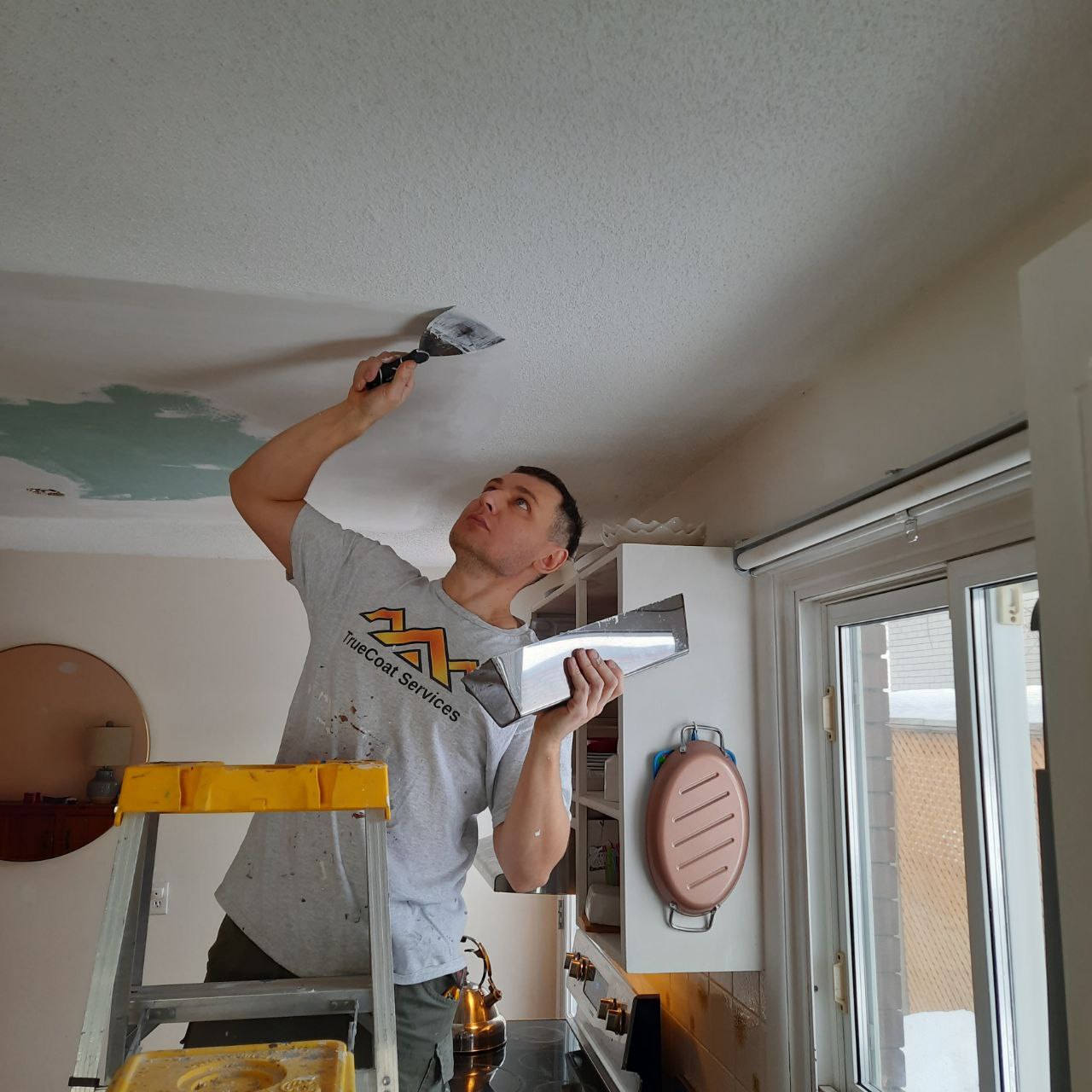TrueCoat Services tradesperson in a branded shirt scraping and skim-coating a damaged ceiling from a step ladder.