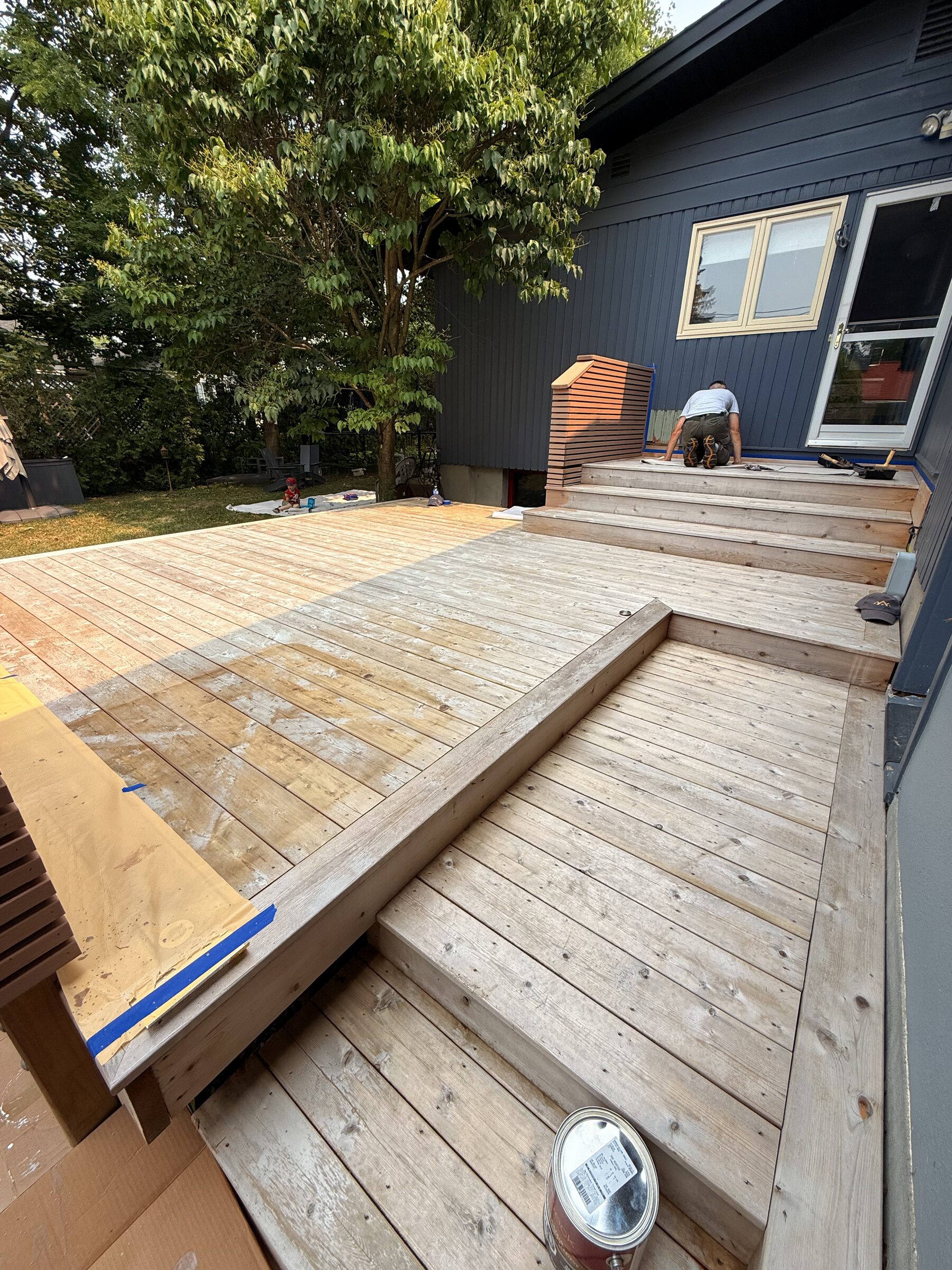 Before: Deck being stained by hand, painter's tape along the edge and a stain can in the foreground