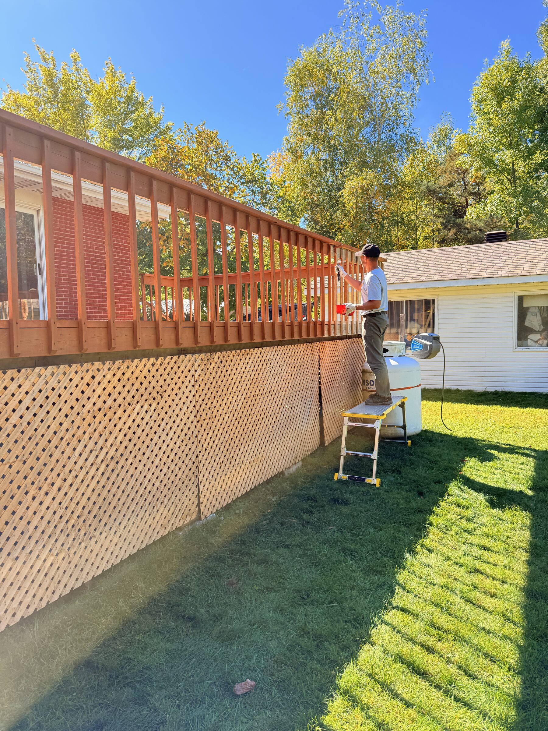 Before: Deck railing being coated by hand beside a red brick wall and diamond lattice skirting