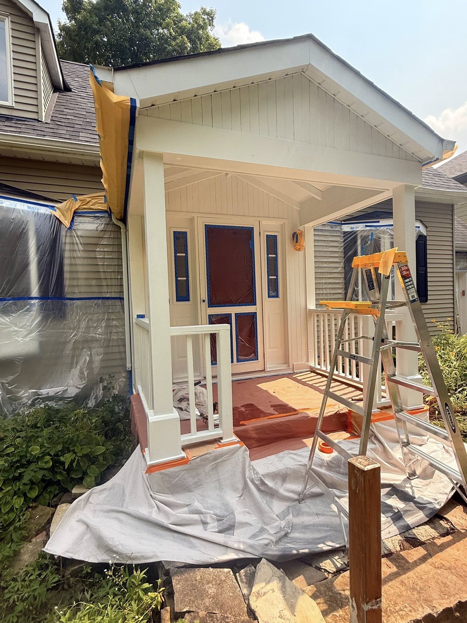 Front porch and siding mid-repaint, with drop sheets over the shrubs, a step ladder and freshly coated cream trim.