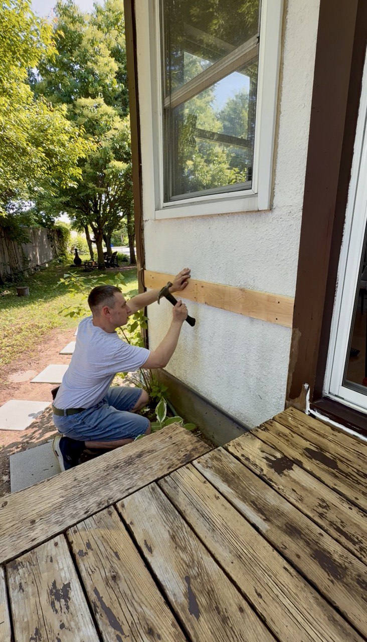 Exterior wall preparation — scraping and sanding stucco siding above a deck before repainting