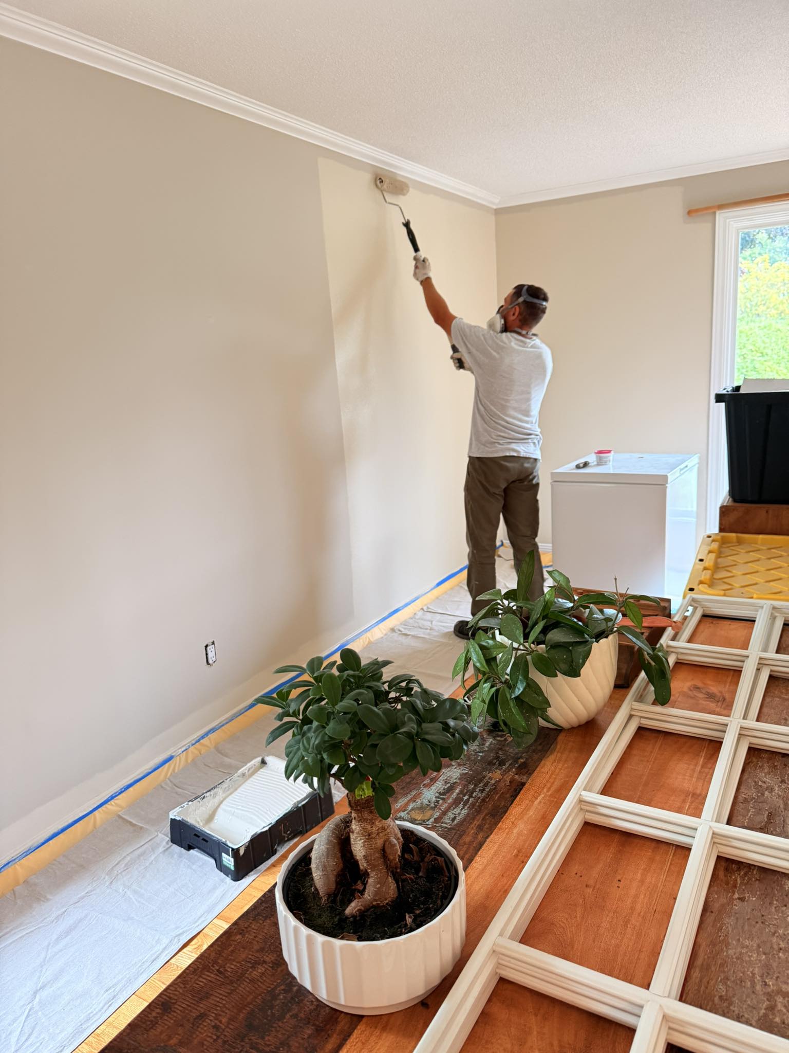 Painter rolling a fresh coat onto an interior wall in a prepped living room, trim removed and floors protected.