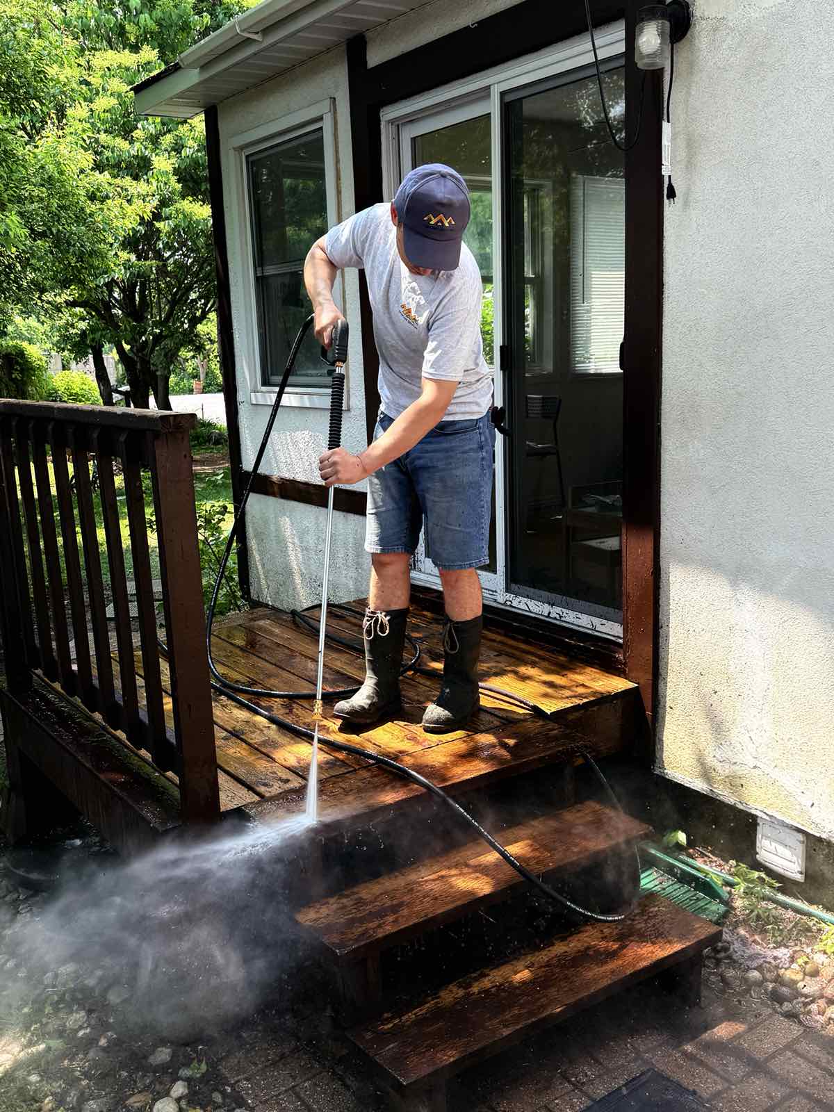 TrueCoat worker pressure washing a weathered wooden deck and stairs, spray visibly lifting dirt from the boards.